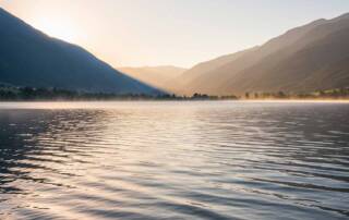 A serene landscape featuring a calm lake with gentle ripples reflecting soft morning light, surrounded by peaceful mountains or hills in soft, muted tones.